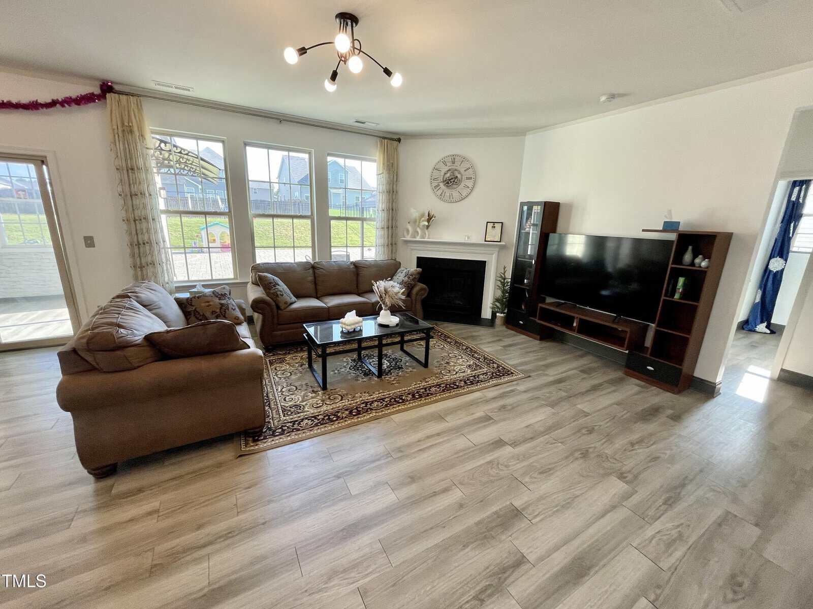 Bright living room with large windows, two brown sofas, a patterned rug, coffee table, fireplace, wall clock, TV, and entertainment unit. Light wood flooring and modern ceiling light fixture complete the space.