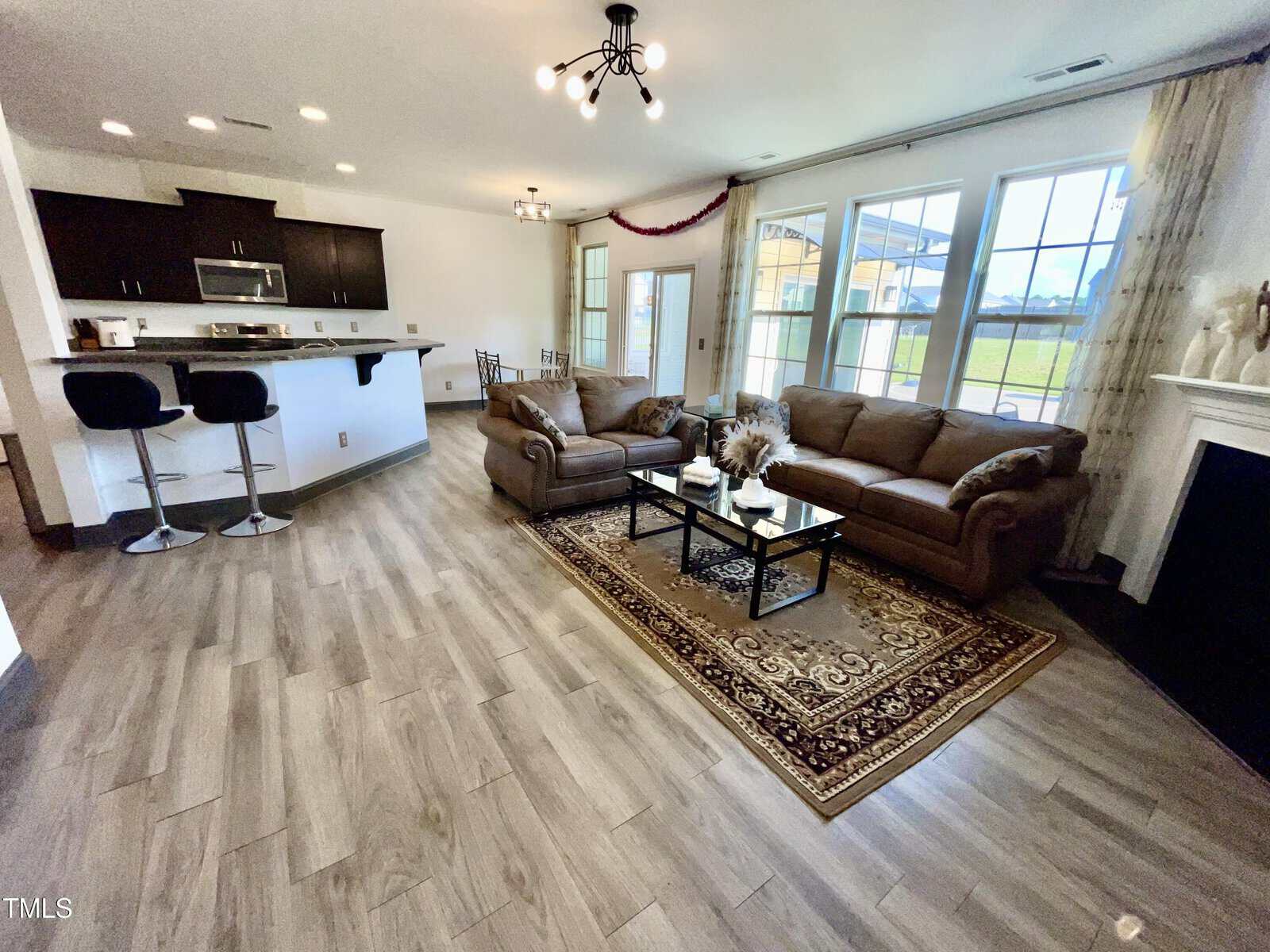 Modern open-concept living room and kitchen with two brown sofas, a rug, a fireplace, large windows, a breakfast bar with stools, and wood flooring. Natural light fills the space.