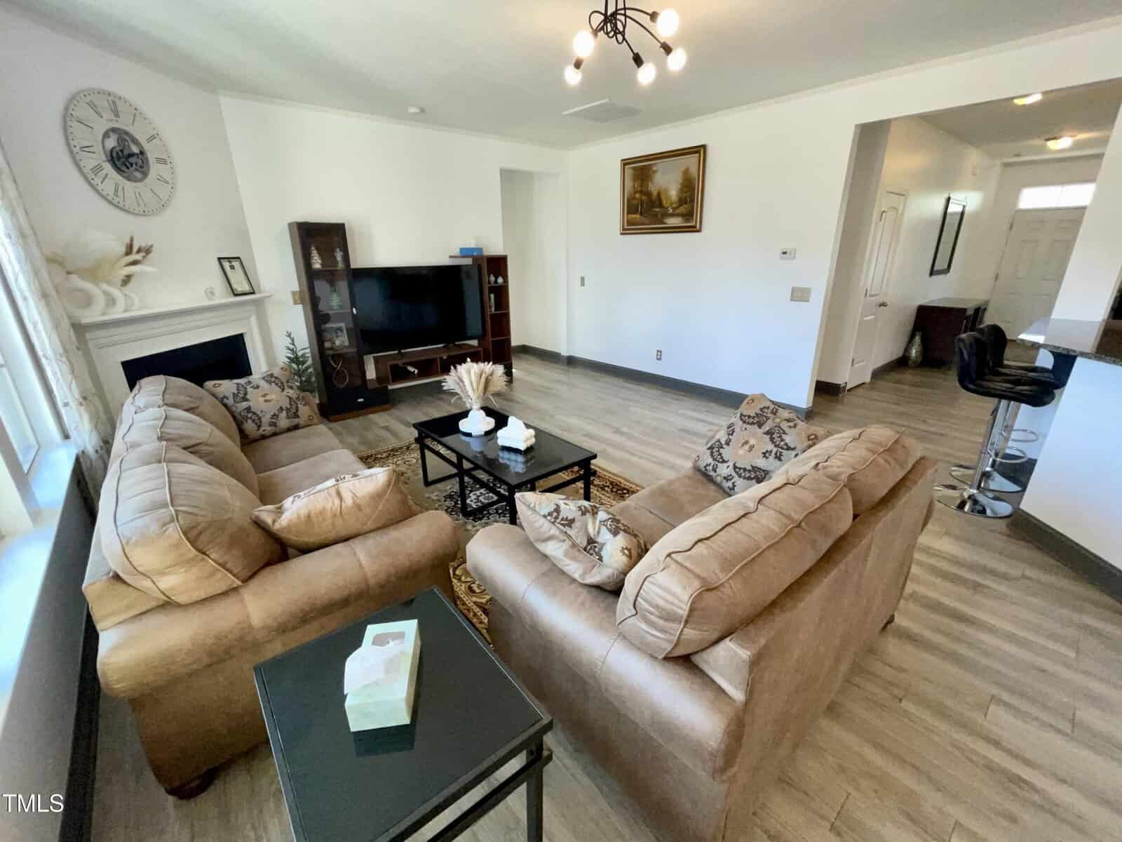 A cozy living room with two tan sofas, a TV on a stand, a modern ceiling light, a fireplace, and a small black side table with tissues. The floor is light wood, and a kitchen is visible in the background.