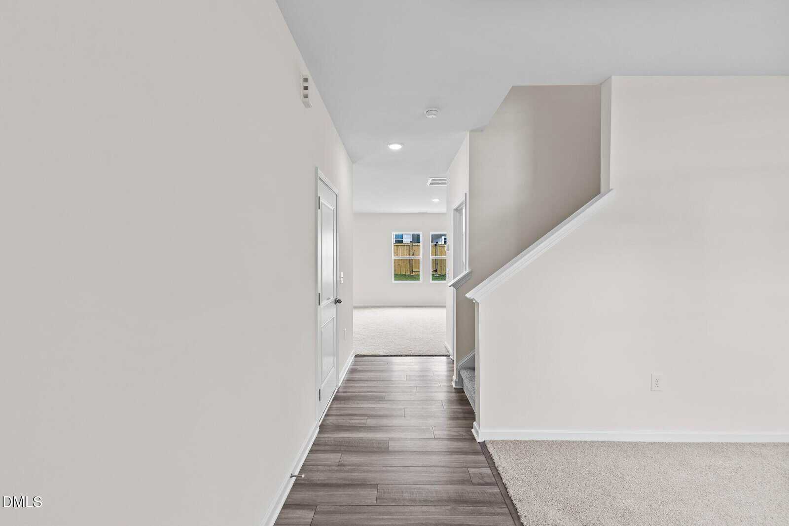 Hallway with light-colored walls and wood flooring leads to a bright living area with large windows. Carpeted stairs are on the right, and a white door is on the left. The space feels clean and modern.