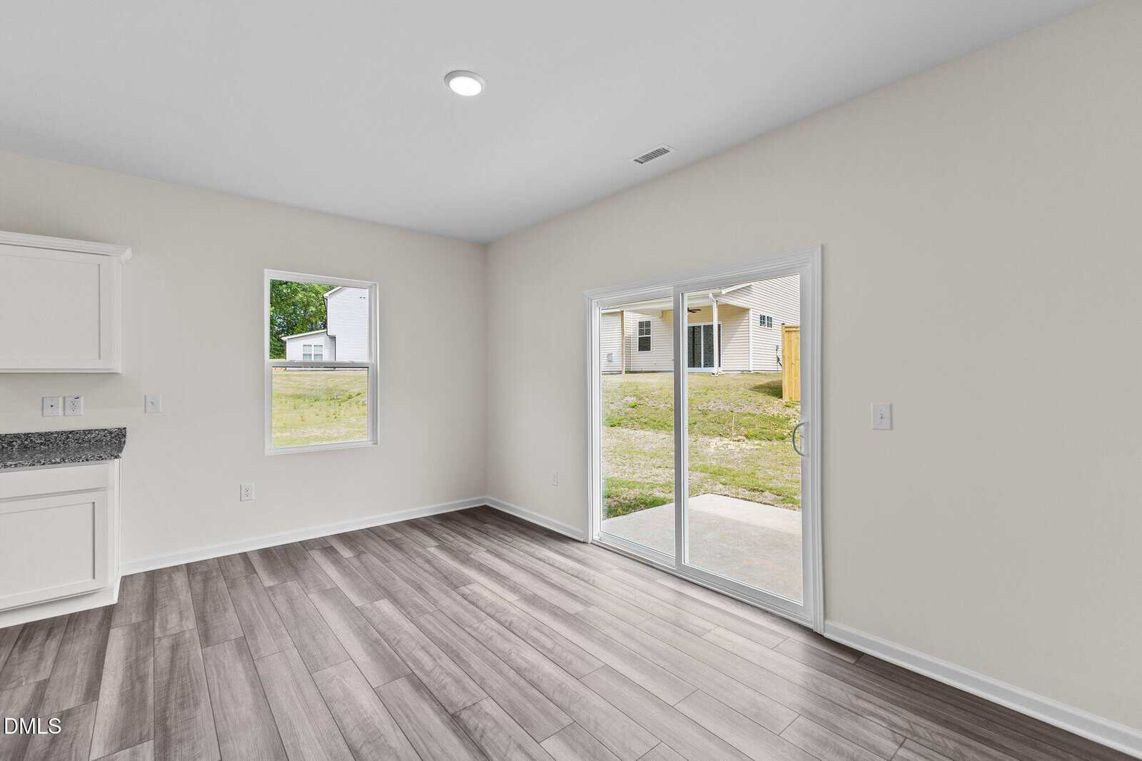 A bright room with light gray wood floors, white walls, a window, and sliding glass doors leading to a backyard with grass and neighboring houses visible outside.