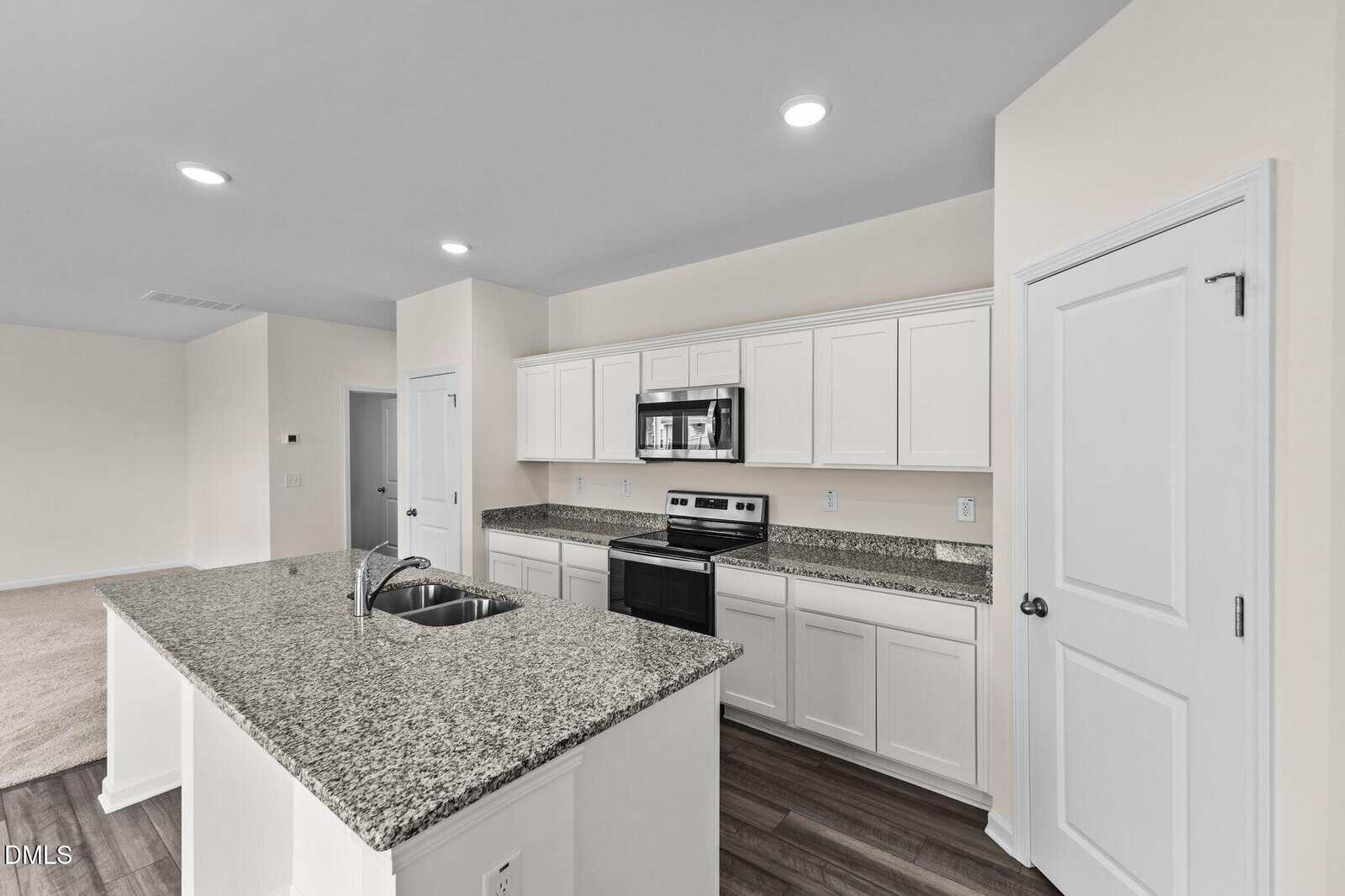 Modern kitchen with white cabinets, granite countertops, stainless steel appliances, island with sink, and light wood floors. The kitchen is open to an adjoining room with neutral walls and recessed lighting.
