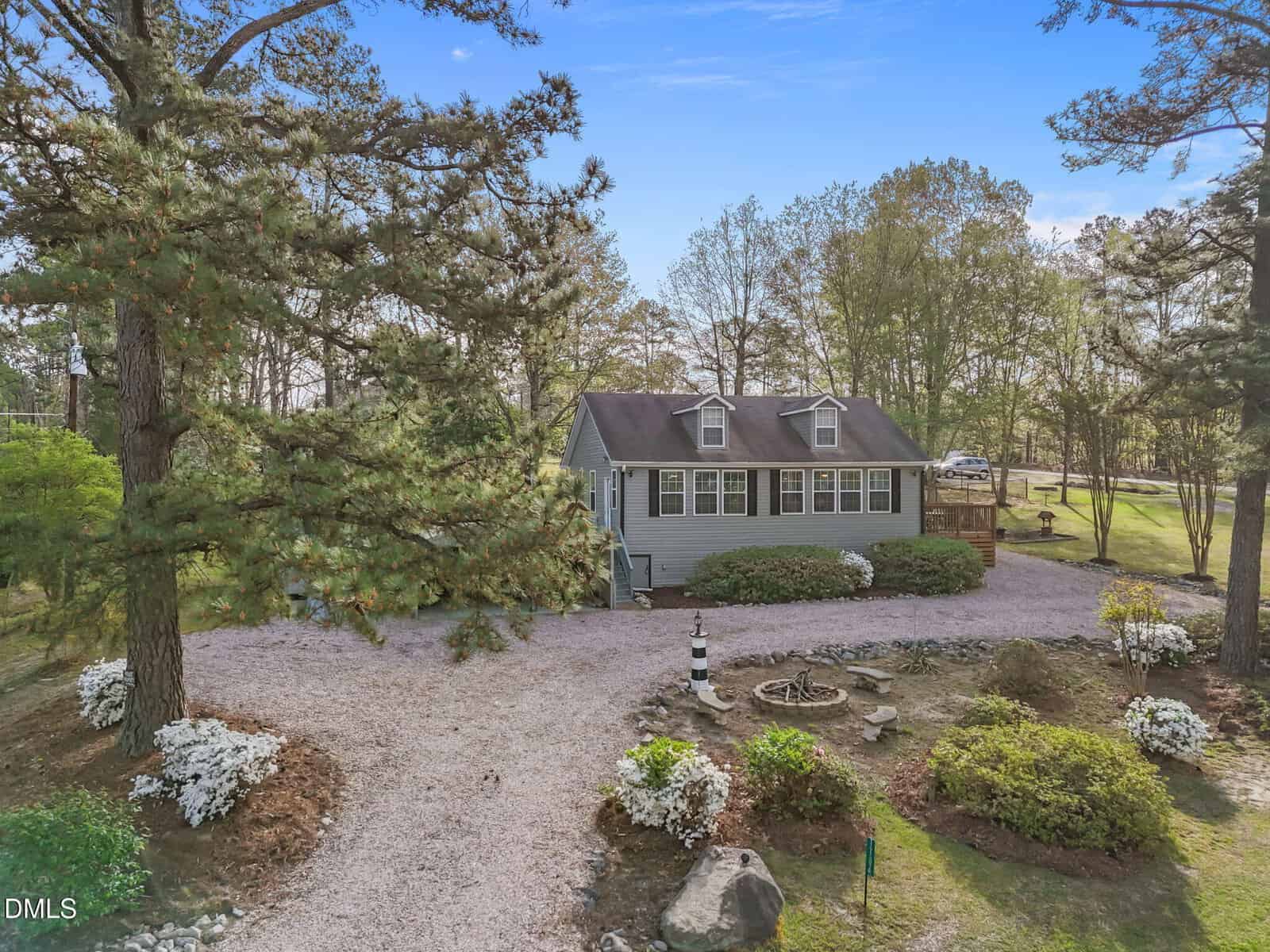 Nestled at 1704 Sagamore Drive in Louisburg, this single-story gray house with charming dormer windows is surrounded by mature trees and beautifully landscaped shrubs. It sits gracefully on a gravel driveway under a clear blue sky, while a small round garden featuring a lighthouse ornament adds character to the inviting foreground.