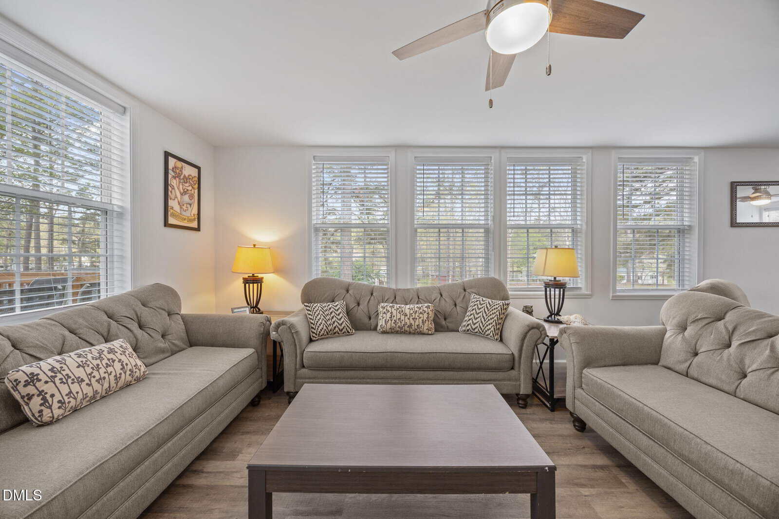 Bright living room with three beige couches, a dark wooden coffee table, two lamps, and large windows with white blinds letting in natural light. Ceiling fan with light is above. Walls are white with minimal decorations.