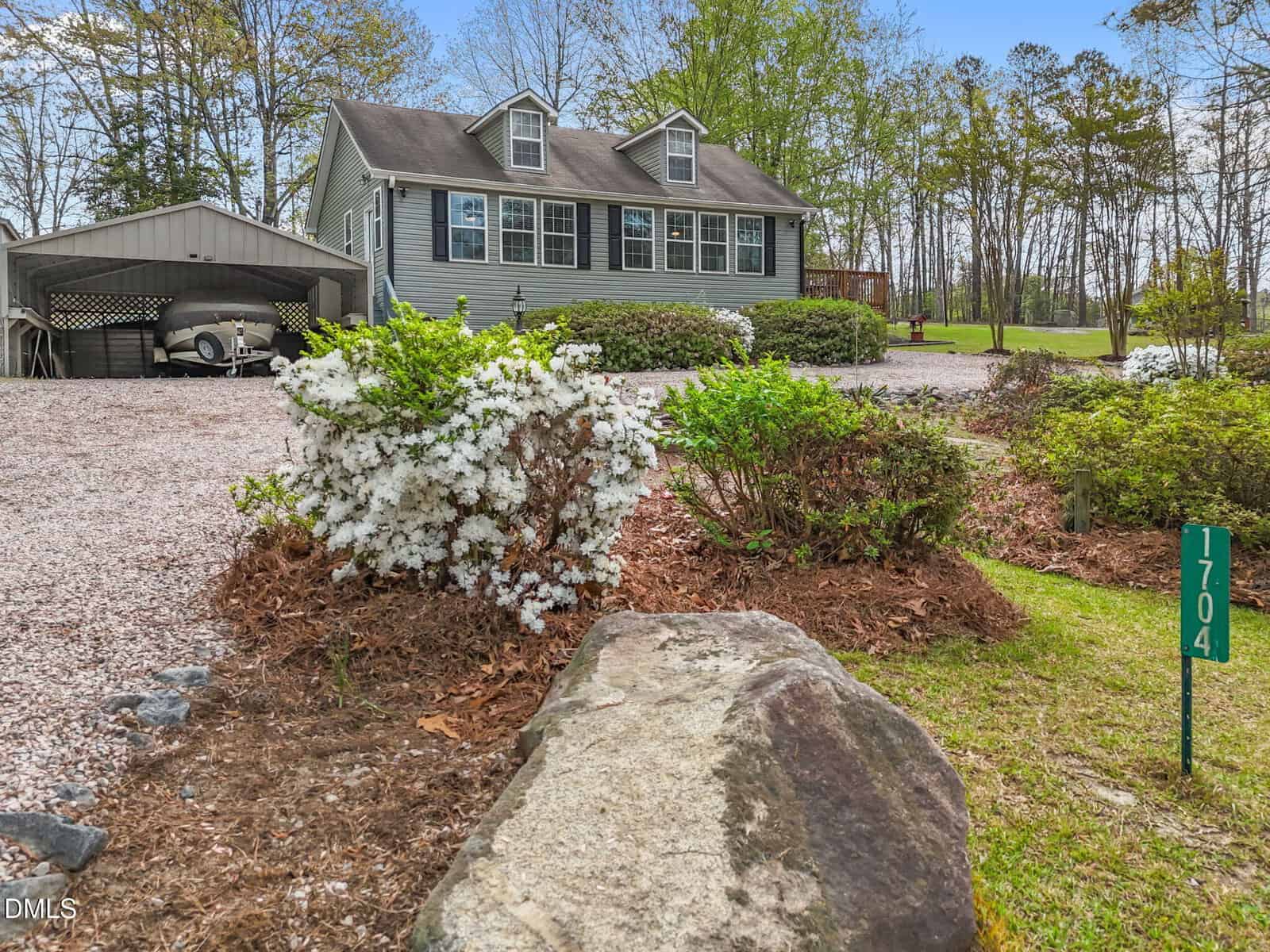 A gray house with dormer windows sits behind a gravel driveway and carport. In the foreground are white flowering bushes, green shrubs, and a green address marker reading “1704.”.
