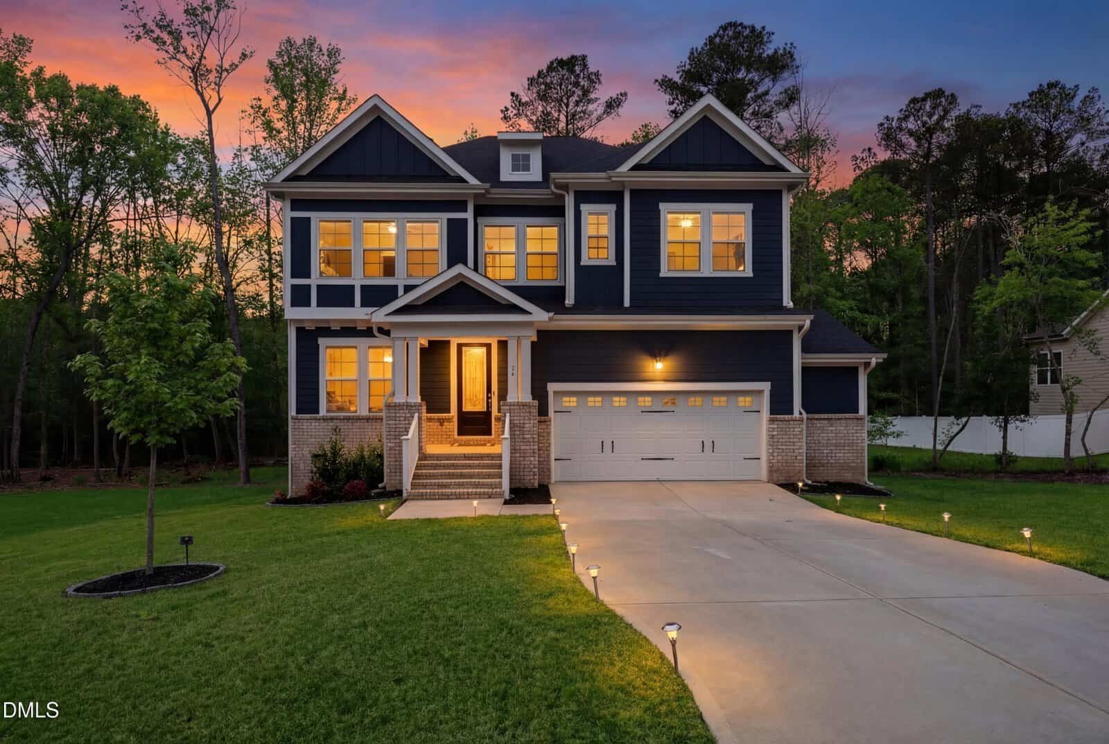 Nestled at 25 Gadwall Court in Zebulon, this two-story navy blue house with white trim features large windows, a front porch, double garage, and a warmly lit exterior at dusk. Garden lights line the walkway, and trees surround its well-kept lawn.