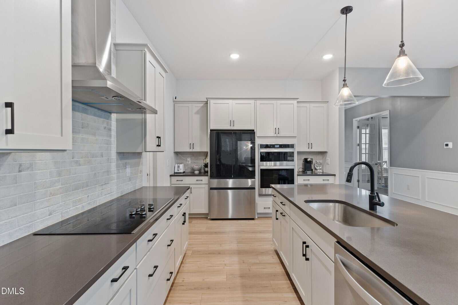 Modern kitchen with light gray cabinets, stainless steel appliances, an island with sink and black faucet, pendant lights, light wood flooring, and a tiled backsplash above a stovetop.