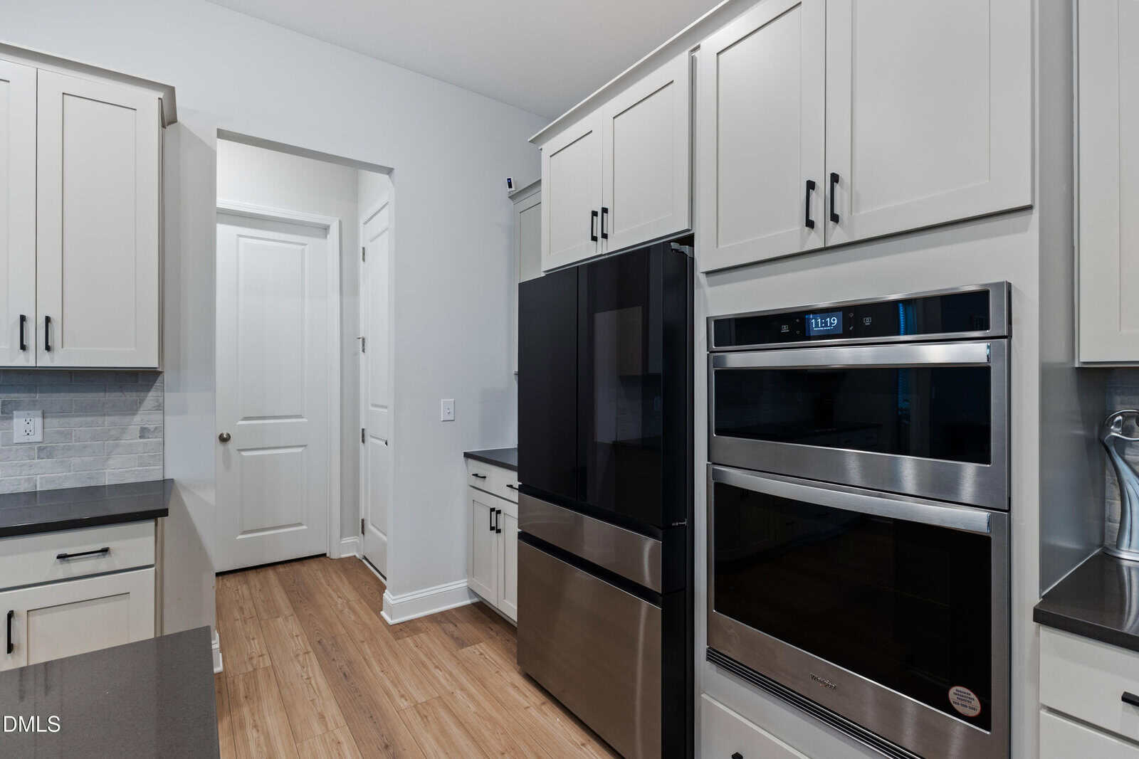 Modern kitchen with white cabinets, black refrigerator, double wall oven, light wood flooring, and gray backsplash. View includes a doorway leading to a hallway with closed white doors.