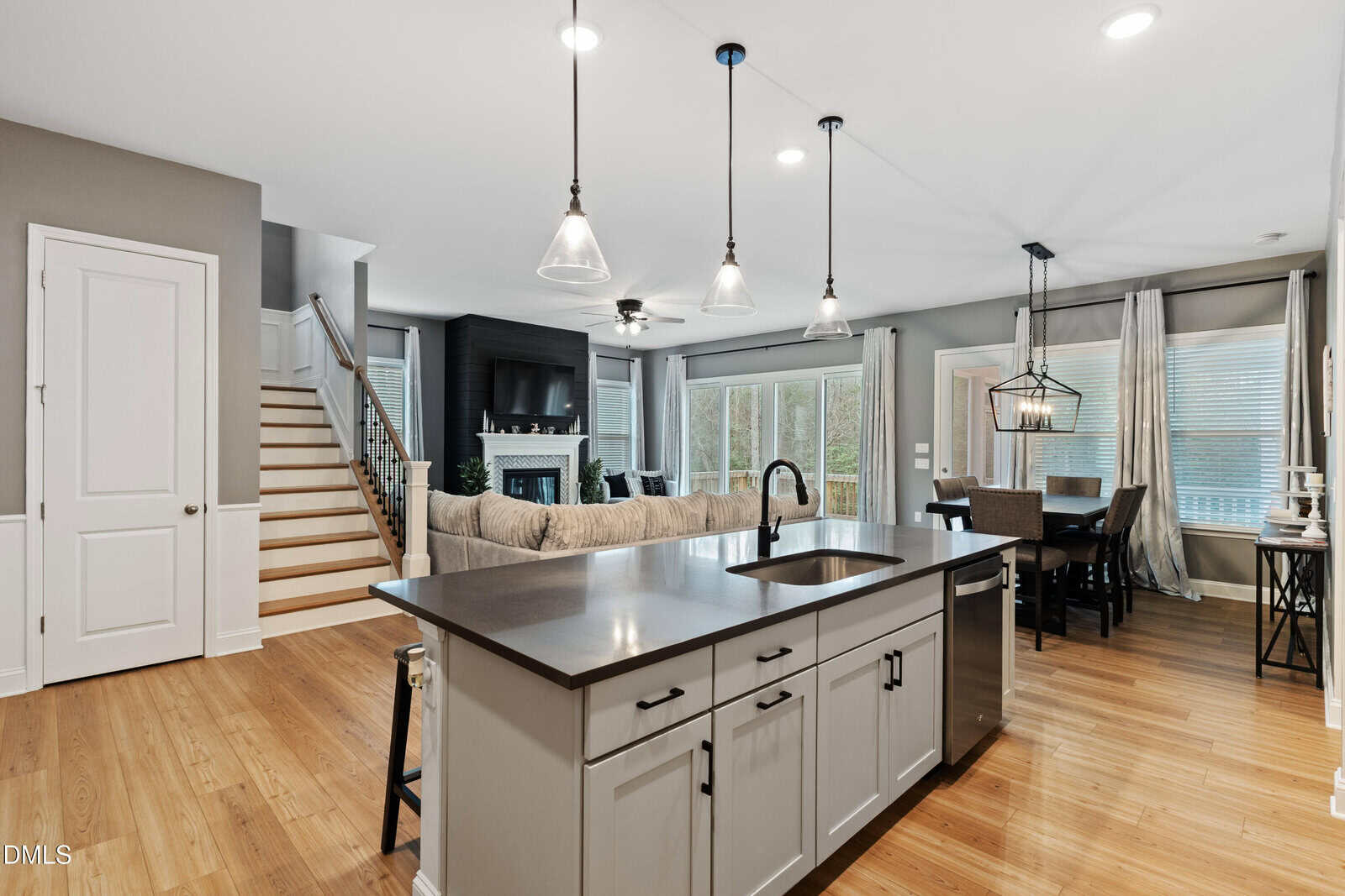 Modern open-concept kitchen and living area with a large island, sink, pendant lights, gray cabinetry, dining table, sectional sofa, fireplace, staircase, and large windows with sheer curtains.