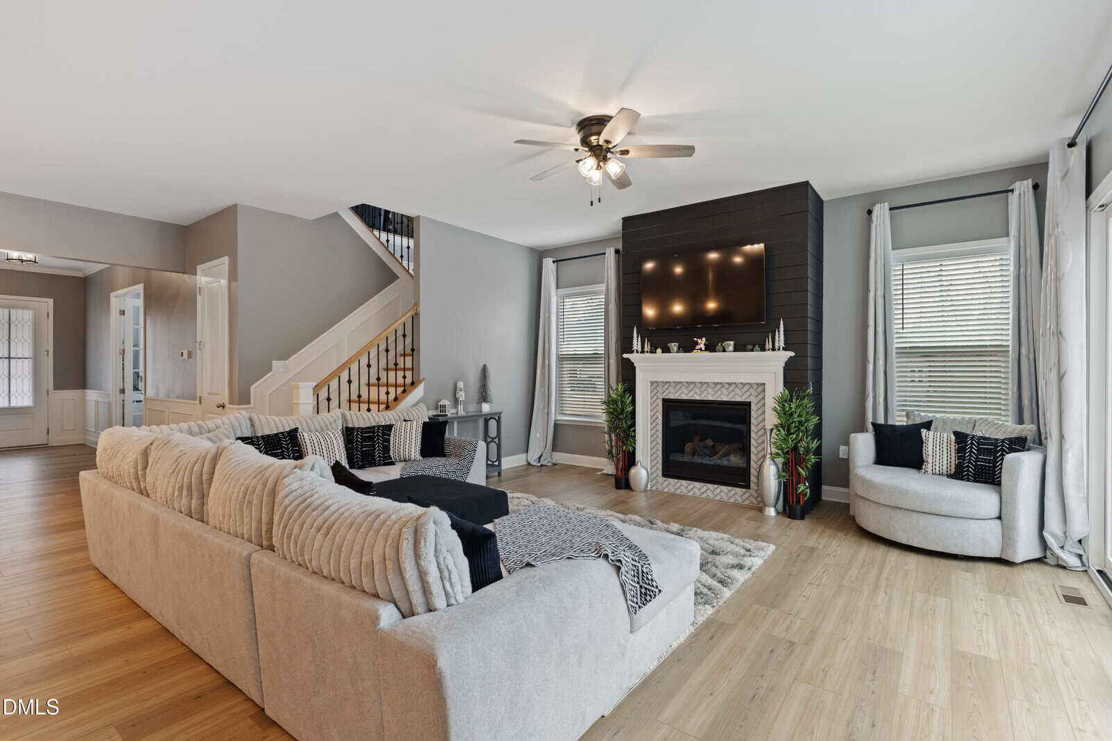 Modern living room with light wood floors, a beige sectional sofa, round chair, fireplace with black accent wall and TV above, and stairs leading to the second floor. Large windows bring in natural light.