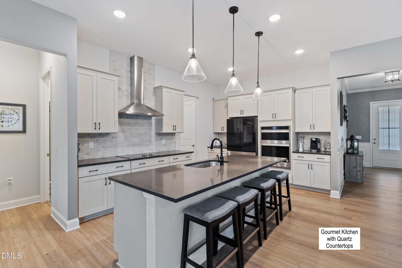 Modern kitchen with white cabinets, stainless steel appliances, a large island with a dark quartz countertop, pendant lights above, and four gray cushioned barstools. A sign reads "Gourmet Kitchen with Quartz Countertops.