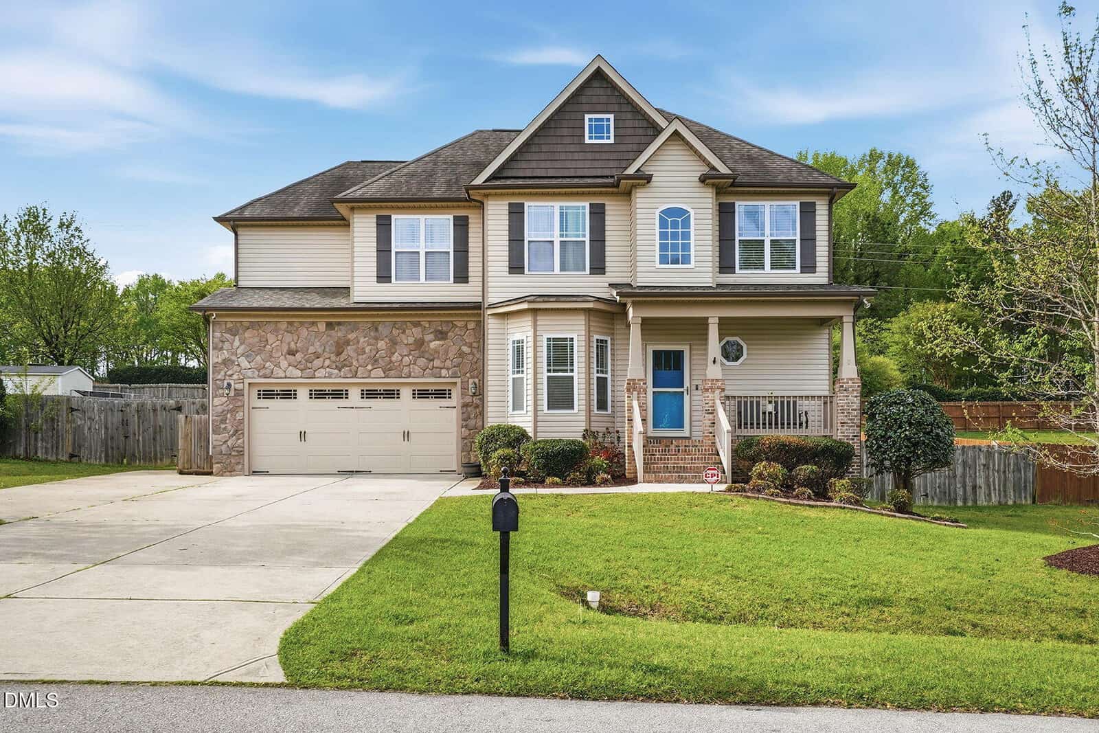 Charming two-story beige house at 25 Cranbrooke Drive, Franklinton, features stone accents, a blue front door, double garage, front porch, manicured lawn, mailbox at the curb, driveway, and trees and fence under a blue sky.
