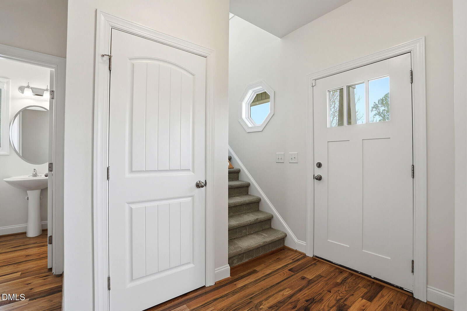 A bright entryway with a white front door, stairs with carpeted steps, a white interior door, hardwood floors, and a small octagonal window on the wall above the stairs. A bathroom sink and mirror are visible to the left.