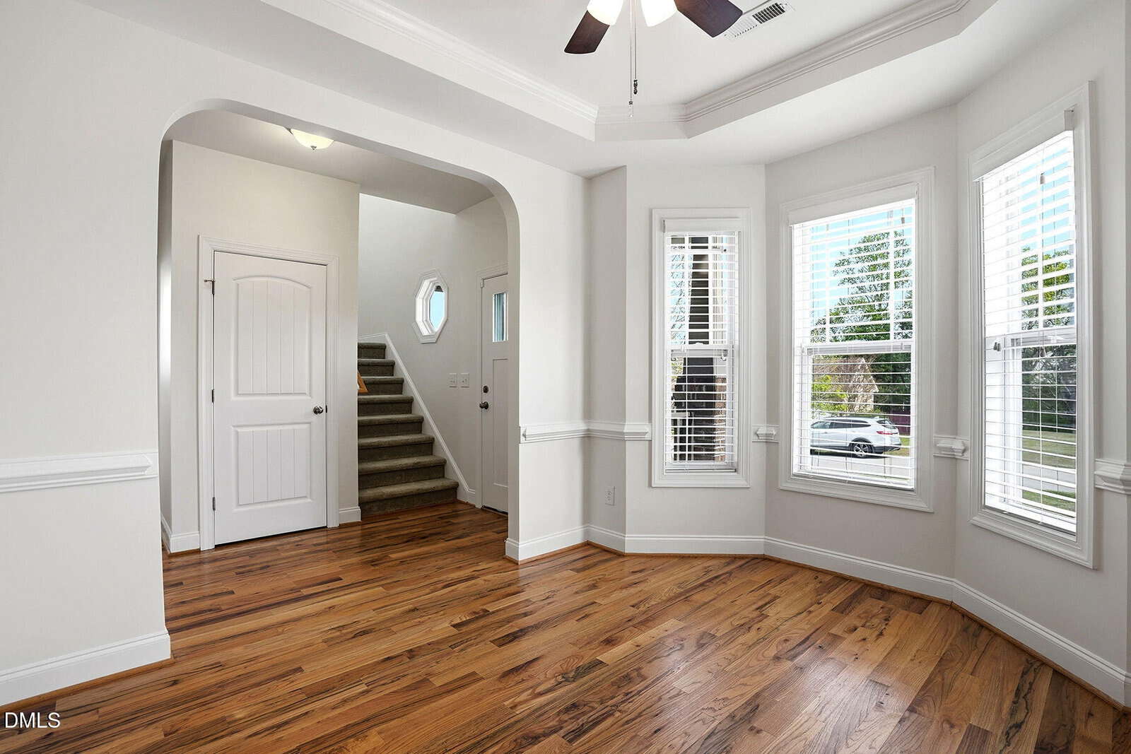 Bright room with hardwood floors, white walls, large bay windows with shutters, a ceiling fan, and an archway leading to a stairway and entry door. Natural light fills the space.