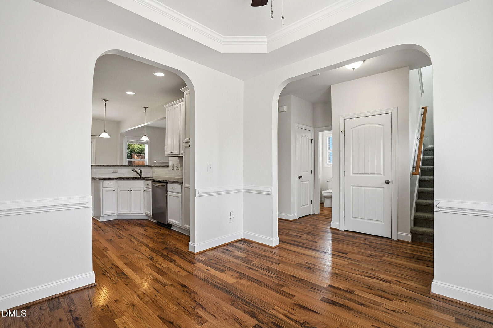 Bright, modern interior with white walls, wood floors, and crown molding. The space includes an open kitchen with pendant lights and stainless appliances, an archway, and a staircase leading upstairs.