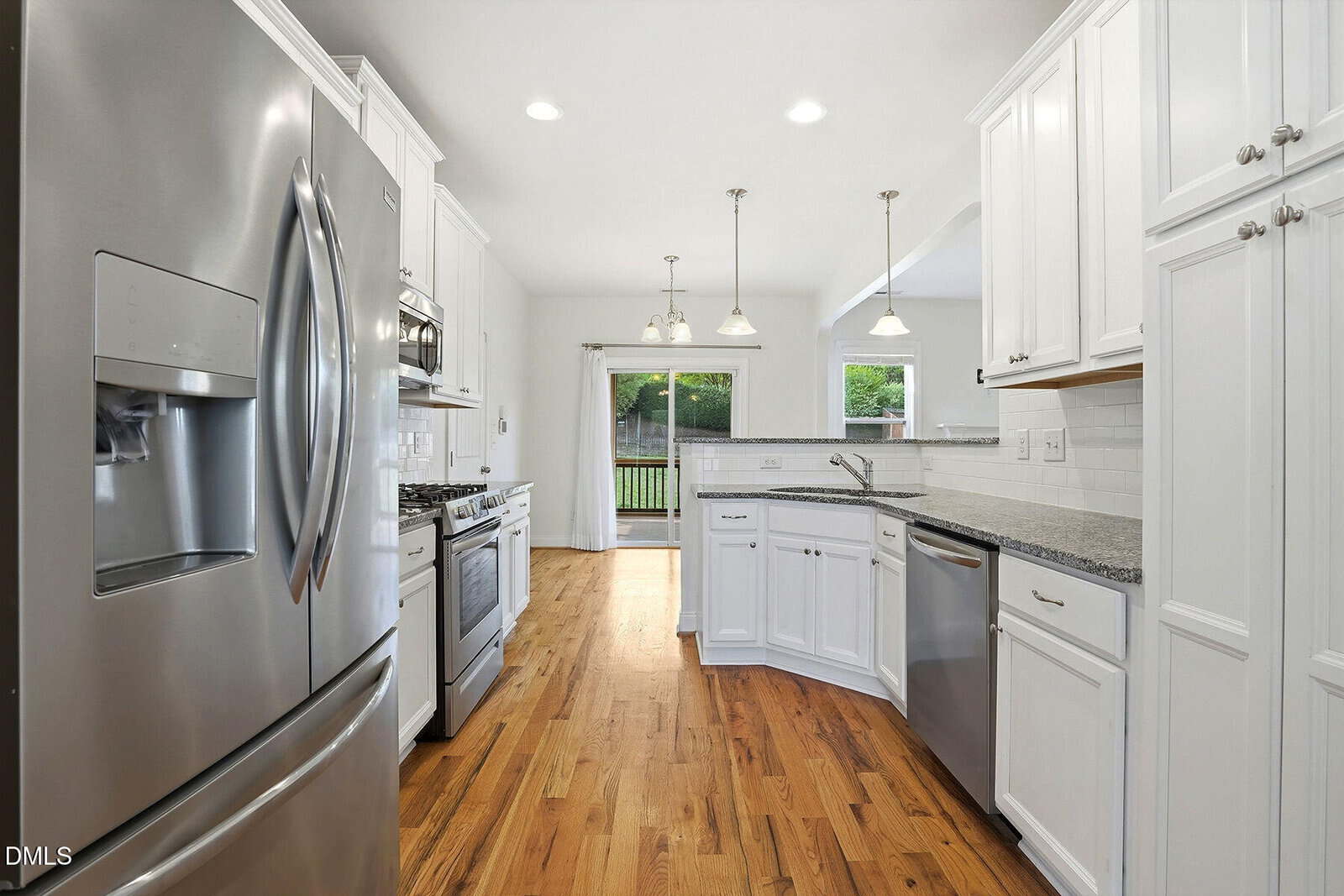 Modern kitchen with stainless steel appliances, white cabinets, granite countertops, hardwood floors, and pendant lights. A sliding glass door at the end opens to a green backyard.