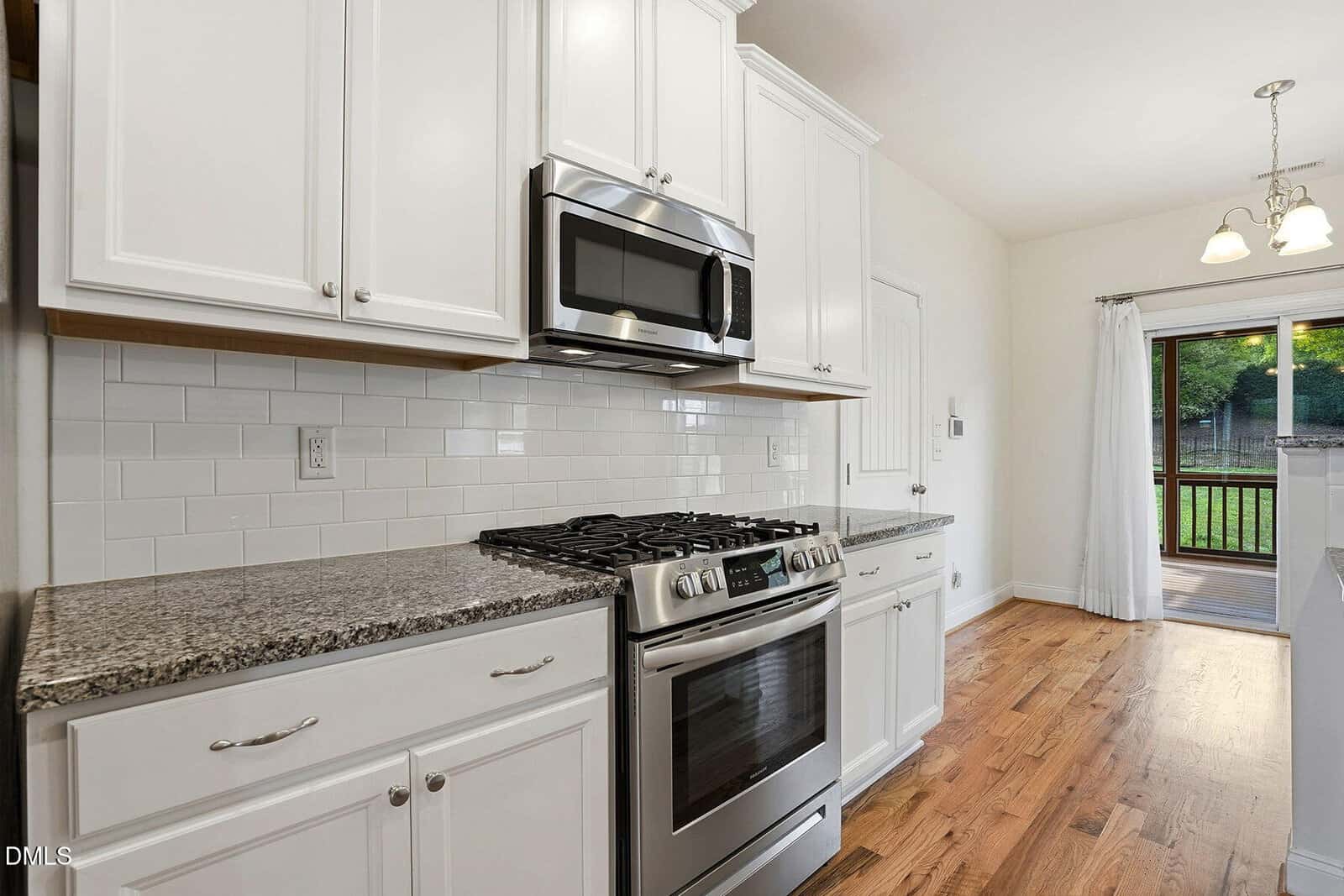 Modern kitchen with white cabinets, granite countertops, stainless steel oven and microwave, subway tile backsplash, hardwood floor, and a glass door leading to a deck outside.