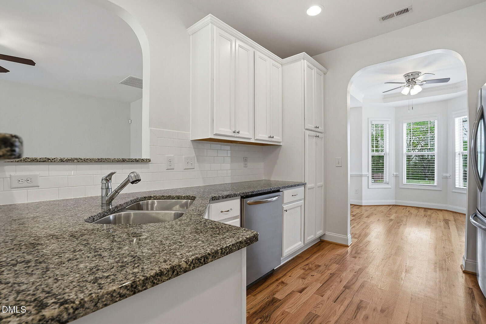 Modern kitchen with white cabinets, granite countertops, stainless steel sink, and dishwasher. Arched doorway leads to a bright dining area with hardwood floors and large windows.