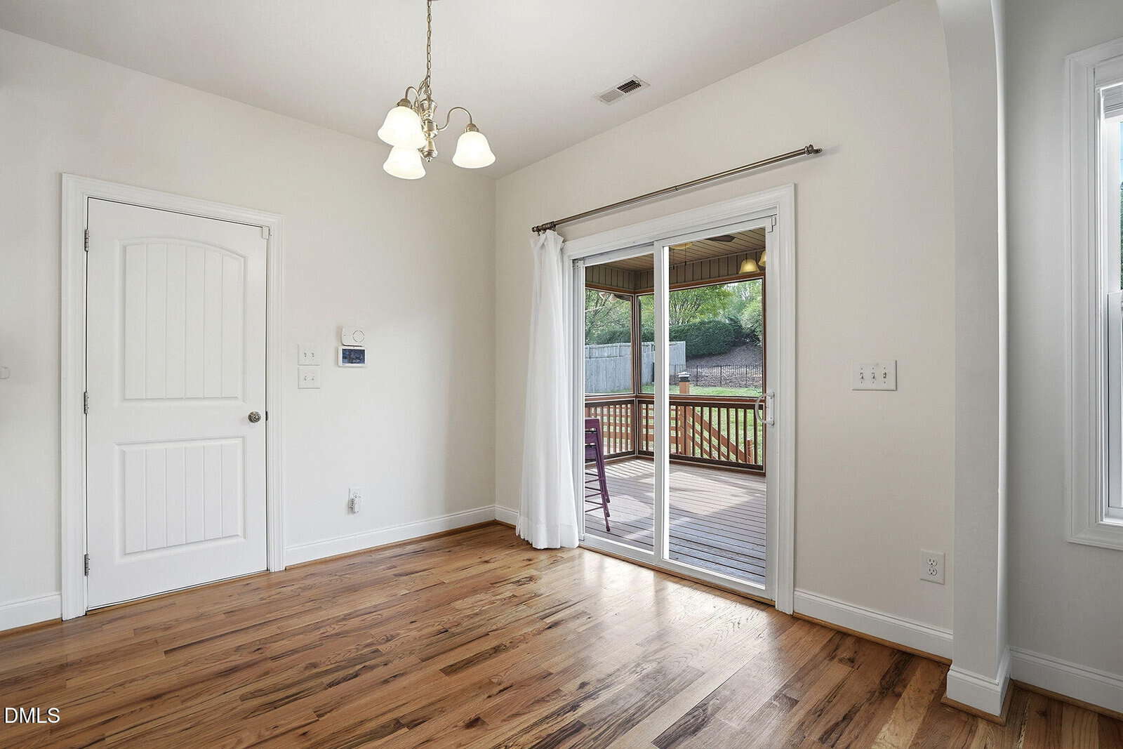A bright, empty room with wooden floors, a white door, and glass sliding doors leading to a wooden deck and fenced backyard. Ceiling light fixture hangs above.