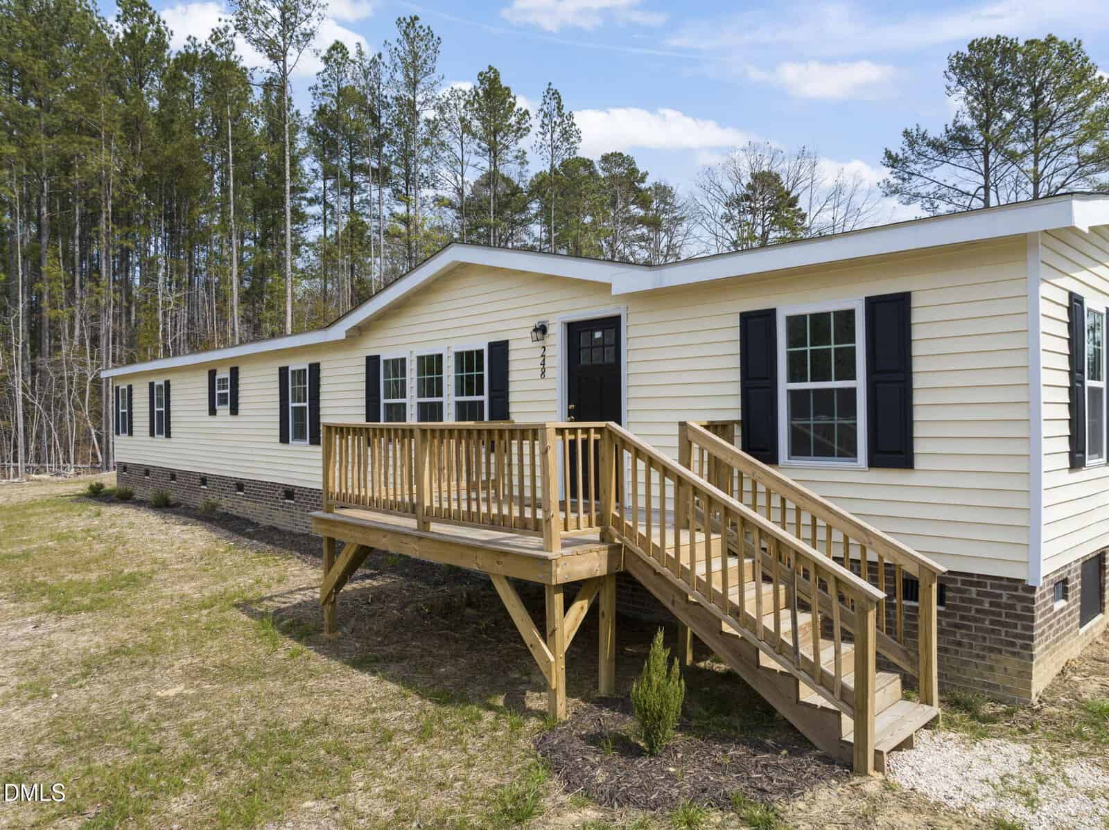 A yellow manufactured home with black shutters and a wooden ramp and stairs leading to the front door, set against a backdrop of tall trees and a grassy yard on 248 Dutchess Lane in Henderson.