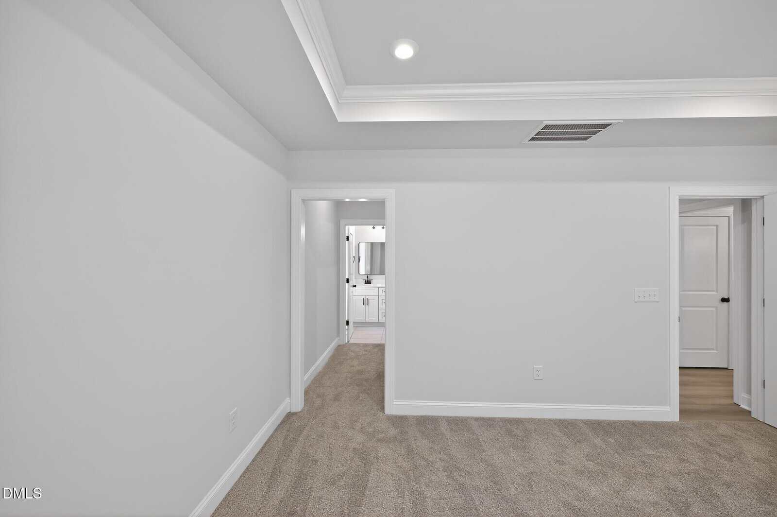 A carpeted room with white walls, tray ceiling, and recessed lighting. Two doorways are visible; one leads to a bright bathroom, and the other to a hallway with a closed white door.
