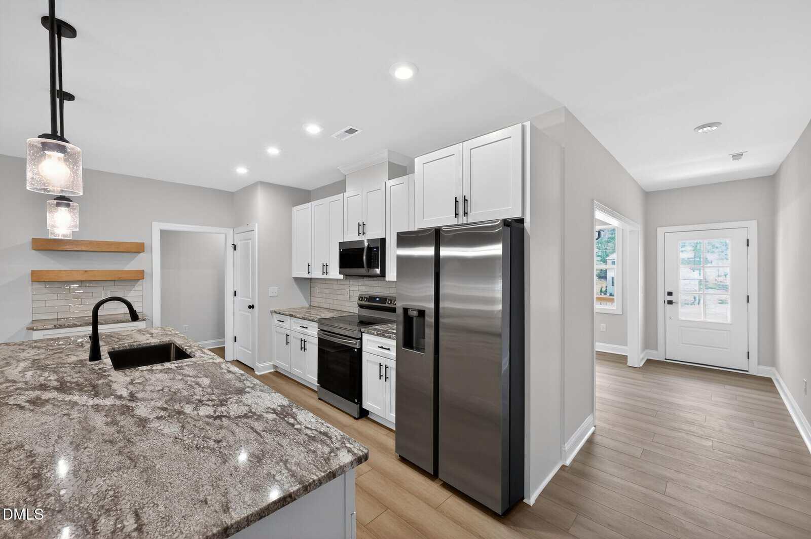 Modern kitchen with granite countertops, stainless steel appliances, white cabinets, wood flooring, open shelves, a farmhouse sink, and neutral gray walls; view includes a doorway leading to a well-lit hallway.