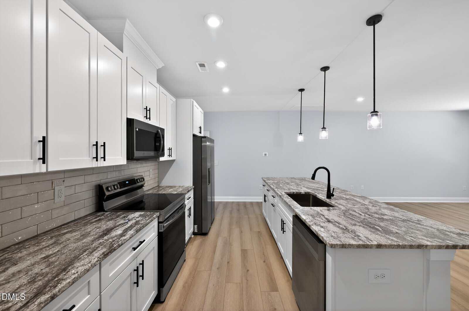 Modern kitchen with white cabinets, stainless steel appliances, gray marble countertops, a large island with a sink, black fixtures, pendant lights, and light wood flooring in an open-plan space.