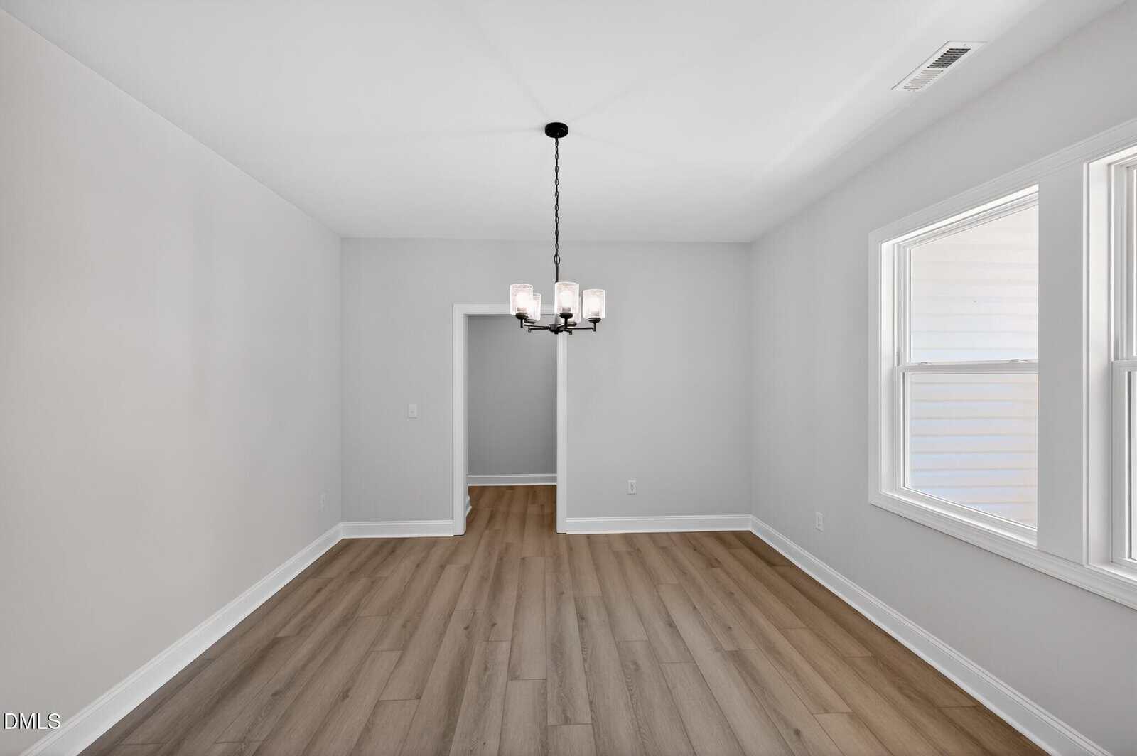 A bright, empty dining room with light gray walls, wood-style flooring, a black chandelier, and two large windows letting in natural light. An open doorway leads to another room.