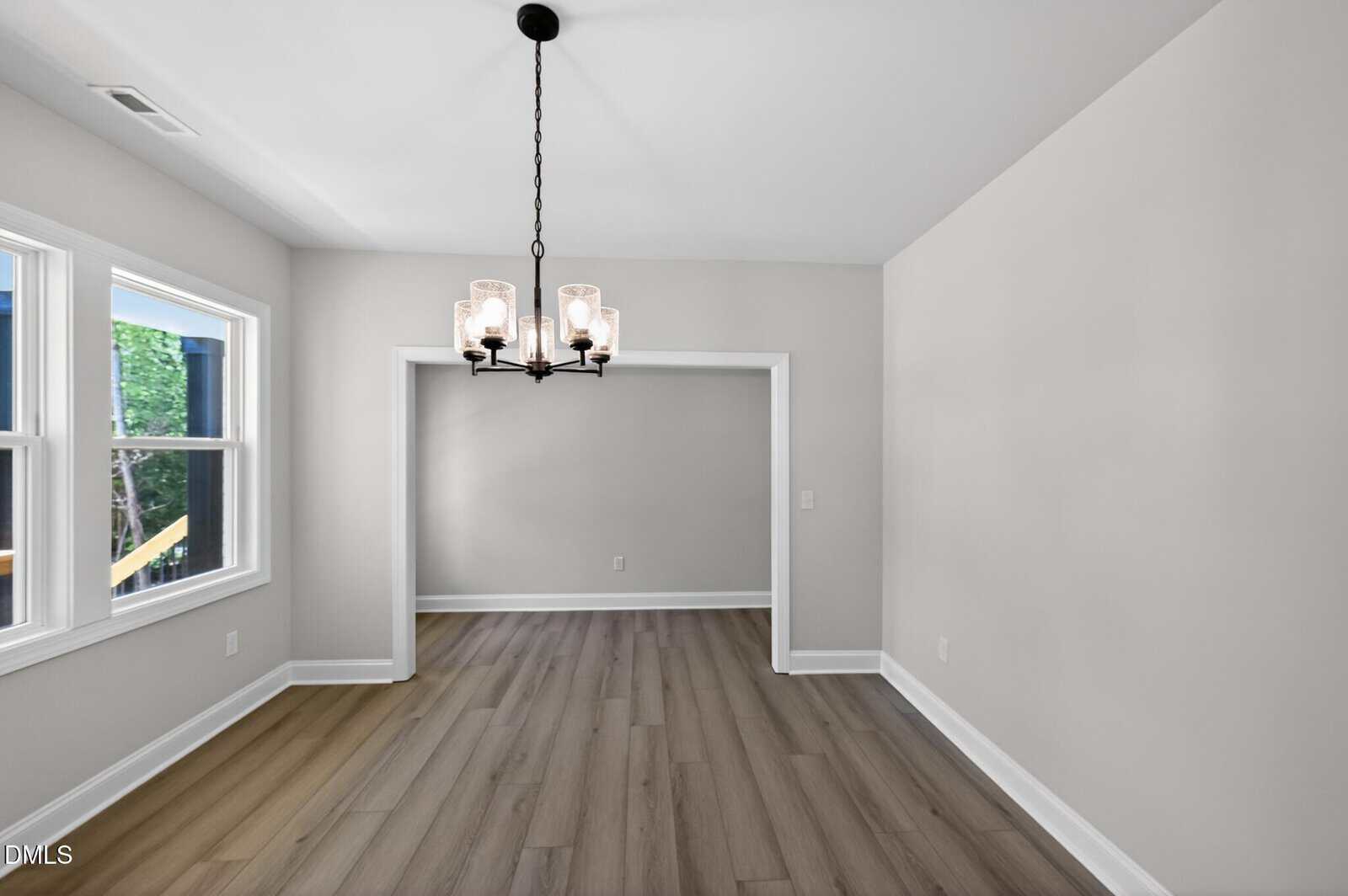 A bright, empty dining room with light gray walls, wood-style flooring, a black chandelier, and large windows letting in natural light. An open doorway leads to an adjacent room.
