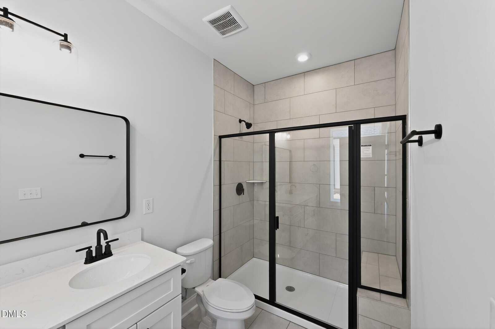 Modern bathroom with light-colored tile and black fixtures, featuring a glass-enclosed shower, white vanity with sink and mirror, and toilet. Towel rack and minimal decor create a clean, contemporary look.