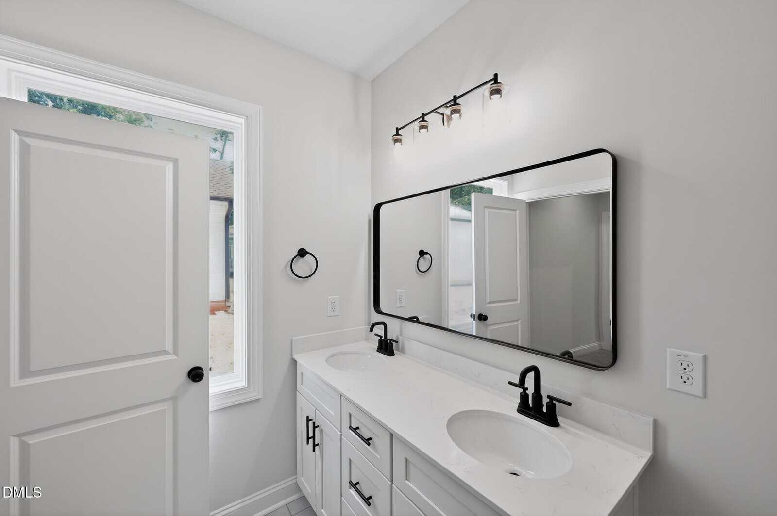 Modern bathroom with a double vanity featuring two sinks, black fixtures, white cabinets, a large rectangular mirror, and three overhead lights; white walls and a window beside an open door.