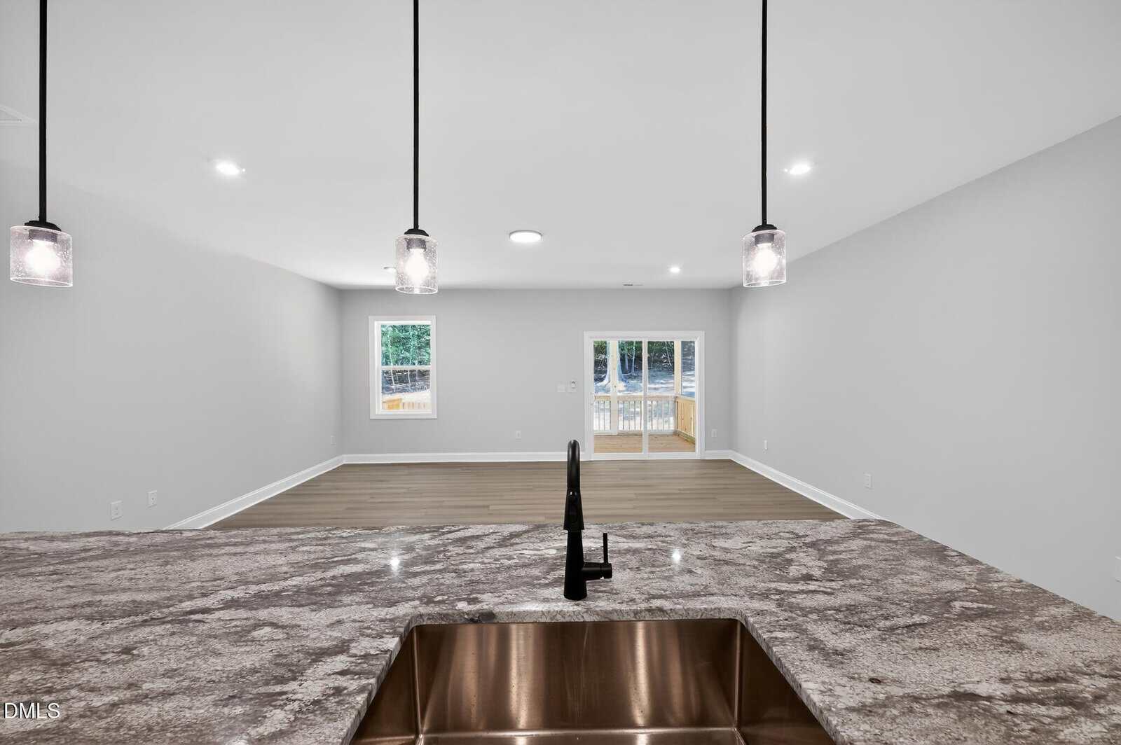 View from a kitchen with a stone countertop and sink, overlooking an open living area with wood floors, white walls, and glass doors leading outside. Three pendant lights hang from the ceiling.
