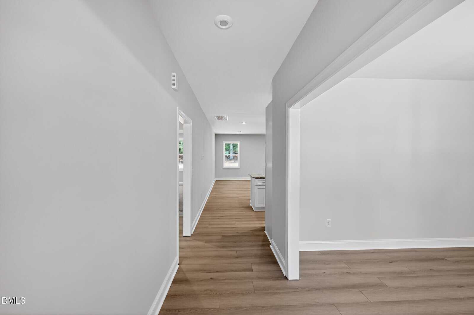 Interior view of a hallway with light gray walls and wood-look laminate flooring. A doorway leads to a room with a window and a kitchen island.