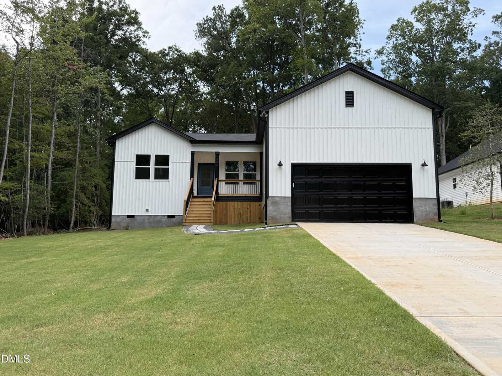 Modern white house with black trim, a large black garage door, front steps, and a wooden porch, set on a green lawn and surrounded by tall trees under a clear sky in the heart of Louisburg at 244 Sequoia Drive.