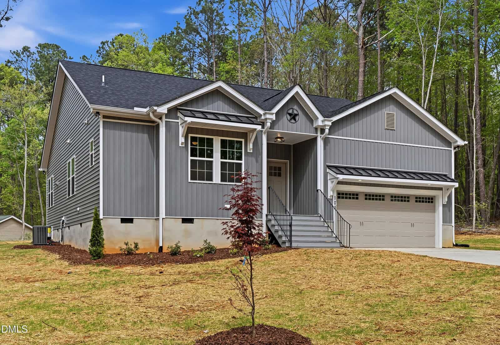 A modern gray house with white trim, front porch steps, a double garage, and landscaped yard with young trees, set against a backdrop of tall green trees and a blue sky.