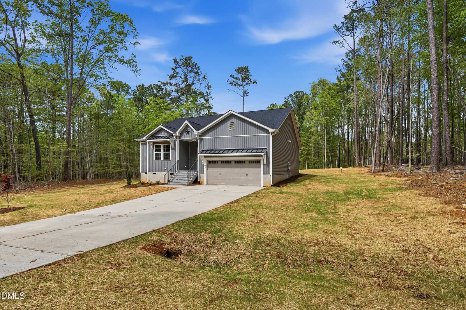 A modern gray house with a two-car garage and gabled roof sits at the end of a long concrete driveway, surrounded by a spacious yard and tall trees under a blue sky.