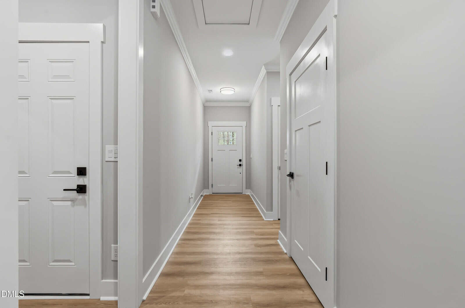 A modern hallway with light gray walls, white doors, and wood flooring, leading to a front door with glass panels. Ceiling lights provide illumination.