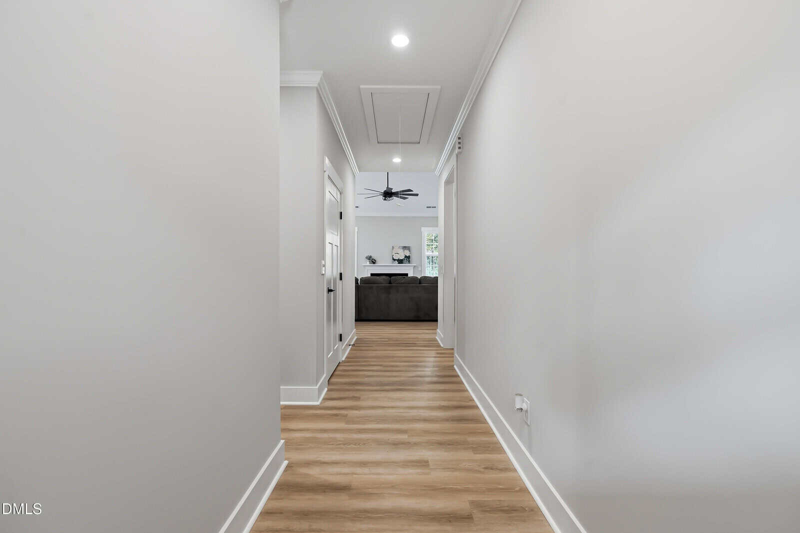 A bright hallway with light gray walls and wood flooring, leading to a living room with a dark sofa, ceiling fan, and large windows letting in natural light.