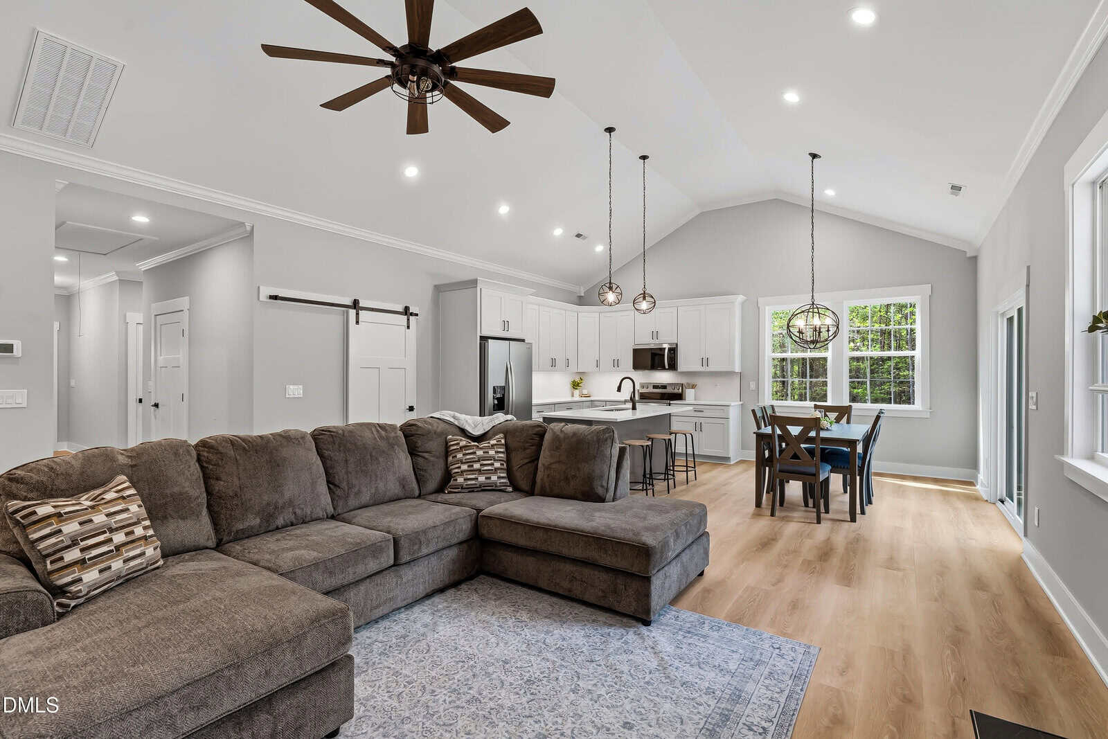 Modern open-concept living room and kitchen with vaulted ceiling, gray sectional sofa, rug, wooden dining table, white cabinets, pendant lights, stainless steel appliances, and large windows letting in natural light.