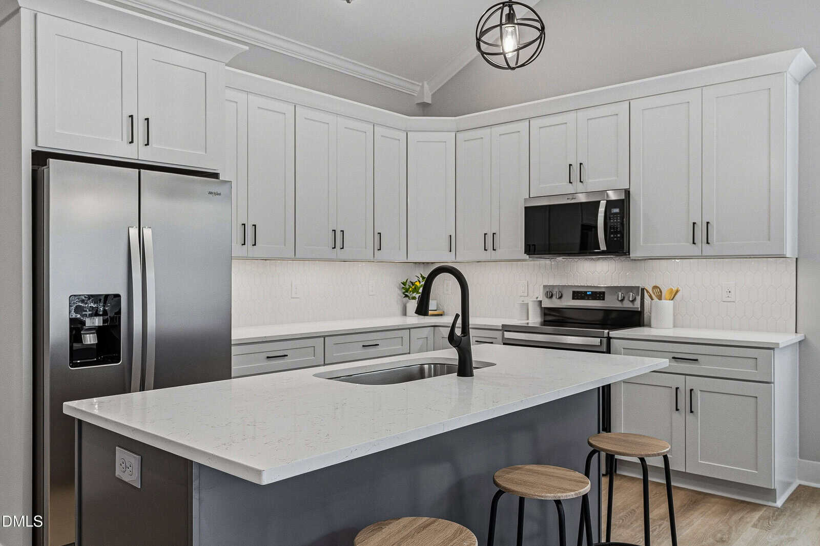 Modern kitchen with white cabinets, stainless steel appliances, an island with a sink and black faucet, three wooden stools, and a round pendant light overhead. The countertop and backsplash are white.