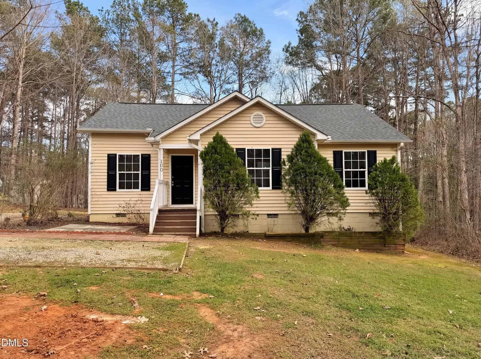 A small, single-story beige house at 100 Mesa Cove, Louisburg, with black shutters, two front-facing windows, and two large bushes by the front steps. There is a patchy lawn, gravel driveway, and tall trees in the background.