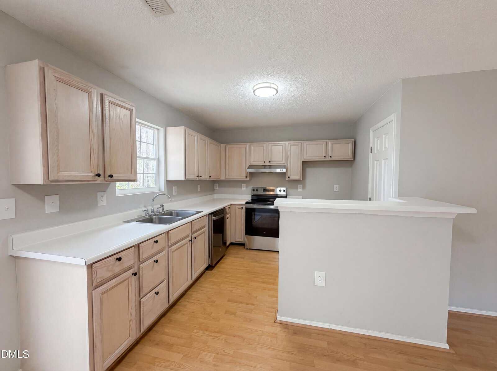 A kitchen with light wood cabinets, white countertops, stainless steel appliances, a double sink under a window, light wood flooring, and pale gray walls. A half wall separates the kitchen from another room.