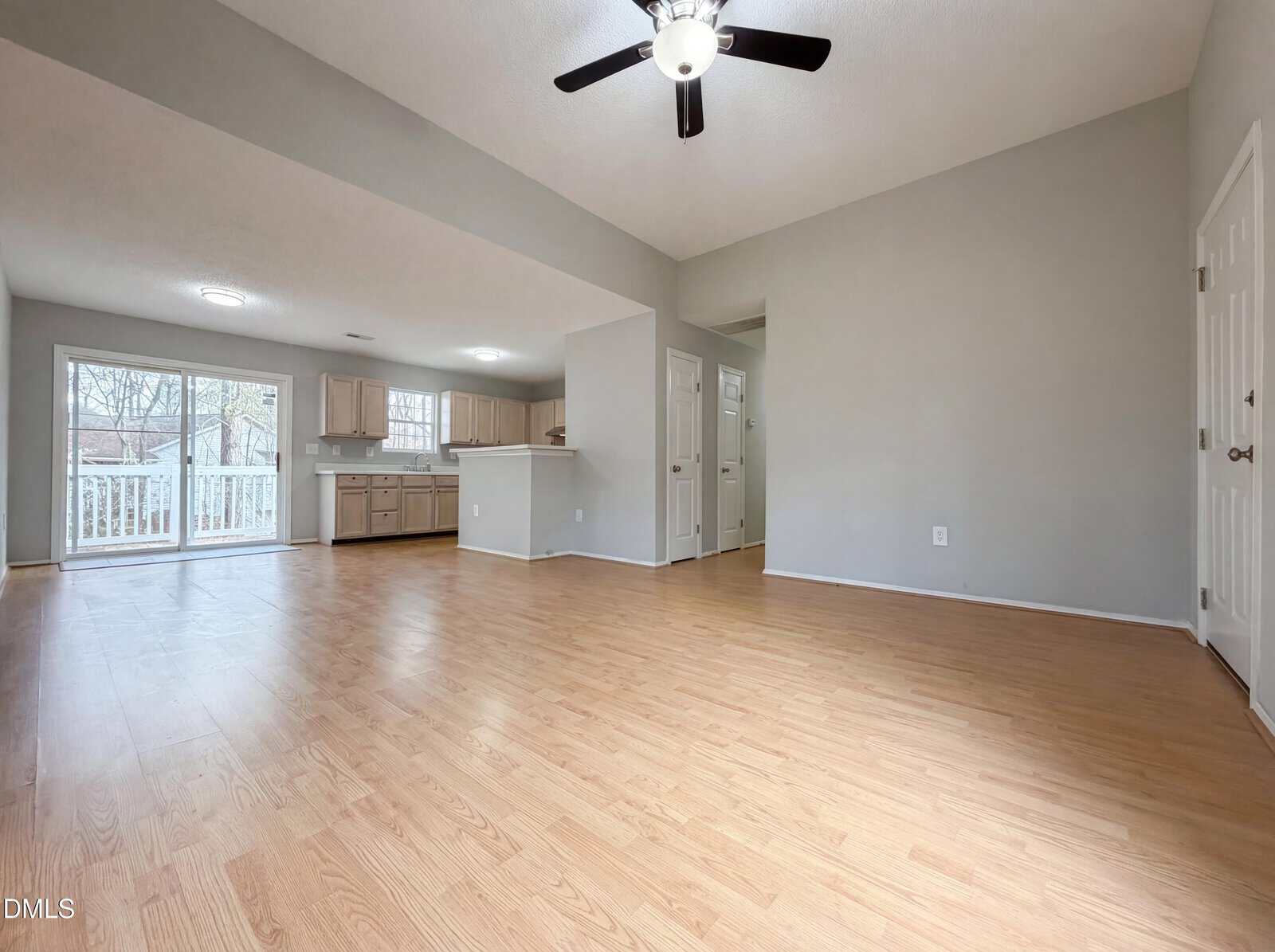 Spacious, empty living area with light wood flooring, neutral walls, ceiling fan, and view into a kitchen with light cabinets. Large sliding glass doors lead to a deck, and several doorways are visible along one wall.