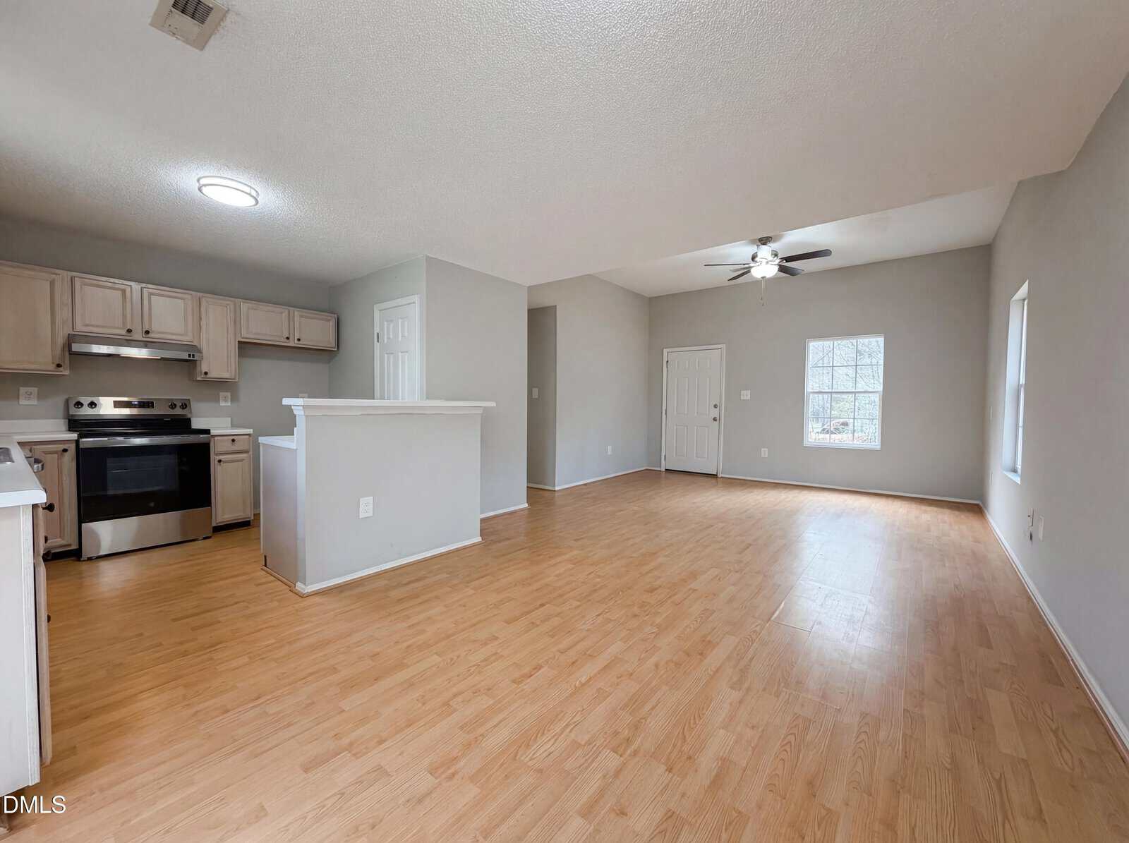 Open-plan kitchen and living room with light wood flooring, neutral walls, white cabinets, stainless steel appliances, ceiling fan, and two large windows letting in natural light.