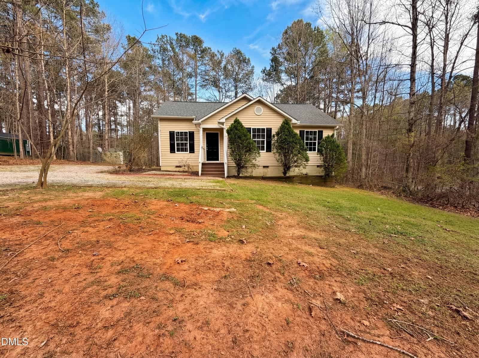 Single-story beige house with white trim and a small porch, surrounded by trees. The front yard has patches of red dirt and green grass. The sky is mostly clear with some clouds.