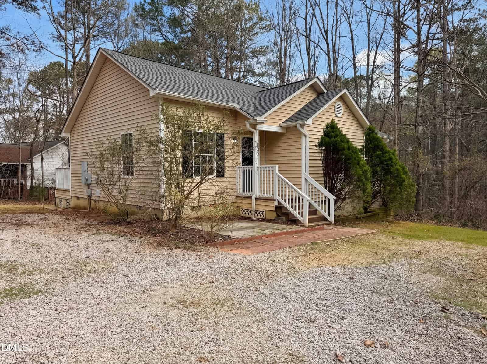 Single-story beige house with white trim, a small front porch with steps, and a gravel driveway. Trees and shrubs surround the property, and the sky is partly cloudy.