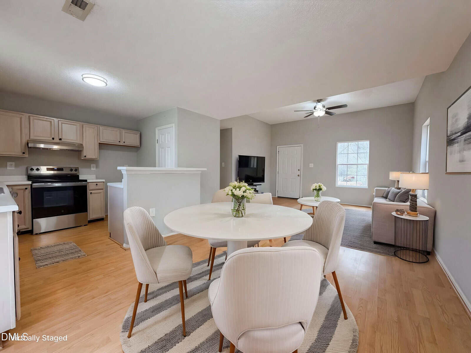 Open-concept living area with a round dining table, four chairs, modern kitchen, cozy living room with a sofa, TV, ceiling fan, and large window. Neutral colors and natural light create a welcoming atmosphere.