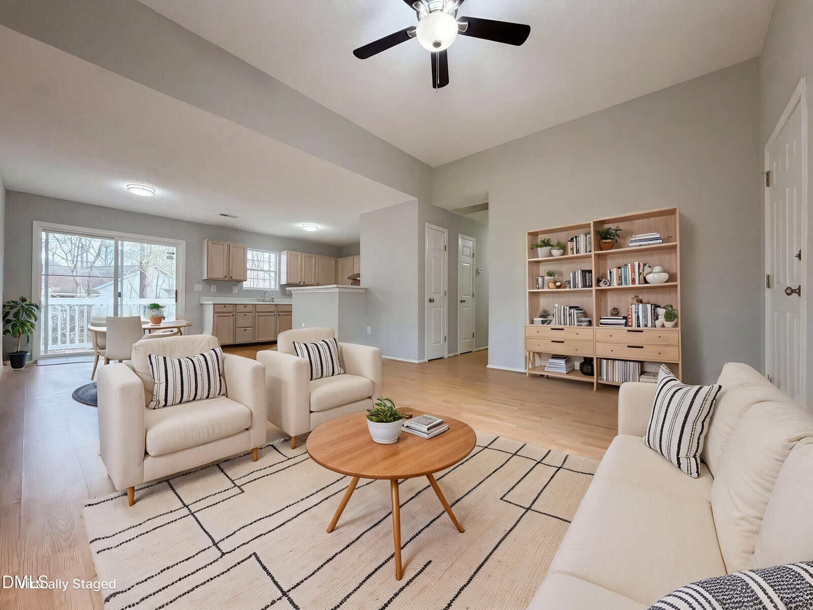 A neutral living room with two beige armchairs, a round wooden coffee table, a patterned rug, bookshelves, and an open view into a kitchen with light cabinets. Large windows and a ceiling fan provide light.