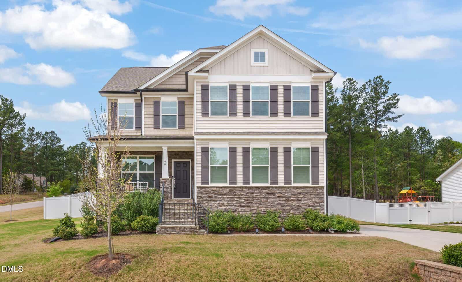 Two-story beige house with stone accents, black shutters, and a front porch nestled at 90 Ironwood Boulevard in Youngsville. The yard is grassy with young trees and shrubbery, while a curved driveway leads to the white fenced backyard. Pine trees and a playground are visible in the background.