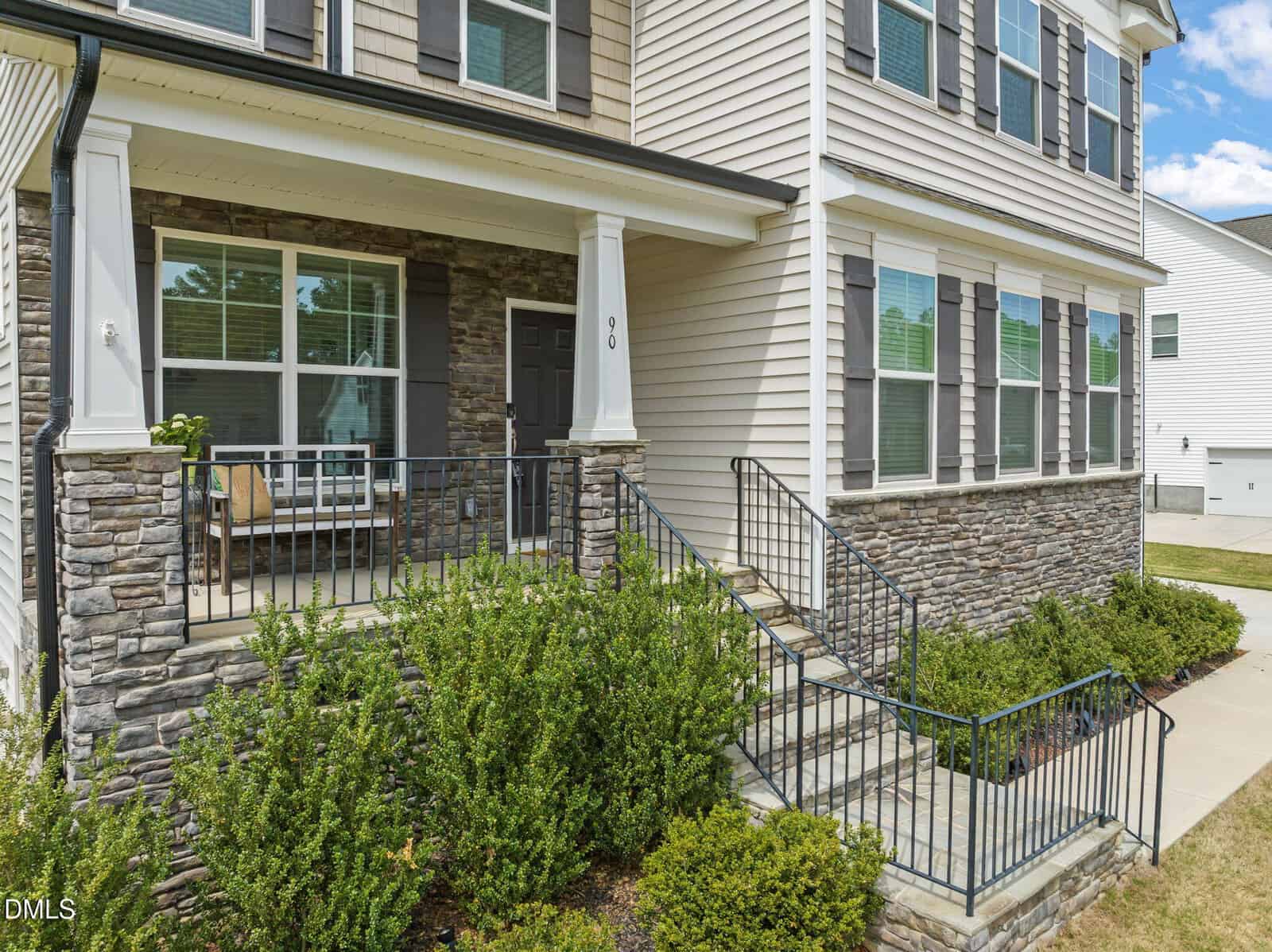 Two-story house with stone and siding exterior, black front door, small porch with seating, metal railing, shrubs along the front, and a concrete walkway on a sunny day.